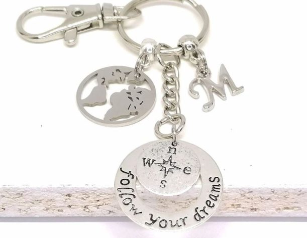 A silver keychian with a world map, the letter M, a compass, and a quote.